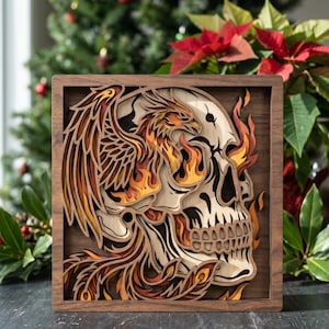 Könnte beinhalten: Ein mehrschichtiges Holz-Kunstwerk, das einen Schädel mit einem Phönix zeigt, der aus Flammen aufsteigt. Das Kunstwerk ist in dunklem Holz gerahmt, wobei der Phönix und die Flammen in Orange-, Gelb- und Brauntönen gehalten sind. Im Hintergrund sind ein Weihnachtsbaum und Poinsettien zu sehen.