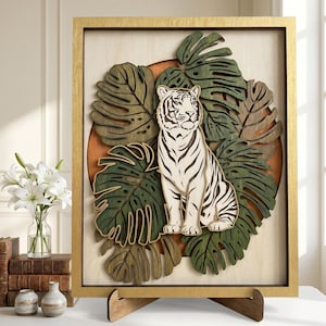 May include: A multi-layered wooden wall art piece featuring a white tiger with black stripes sitting amidst several green monstera leaves. The artwork is set against a circular orange background within a rectangular gold-coloured wooden frame.