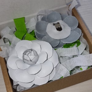 May include: Two white leather flower brooches in a brown box with white tissue paper. One flower is a simple design and the other has a center with a small gray bear.