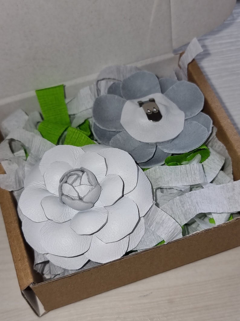May include: Two white leather flower brooches in a brown box with white tissue paper. One flower is a simple design and the other has a center with a small gray bear.
