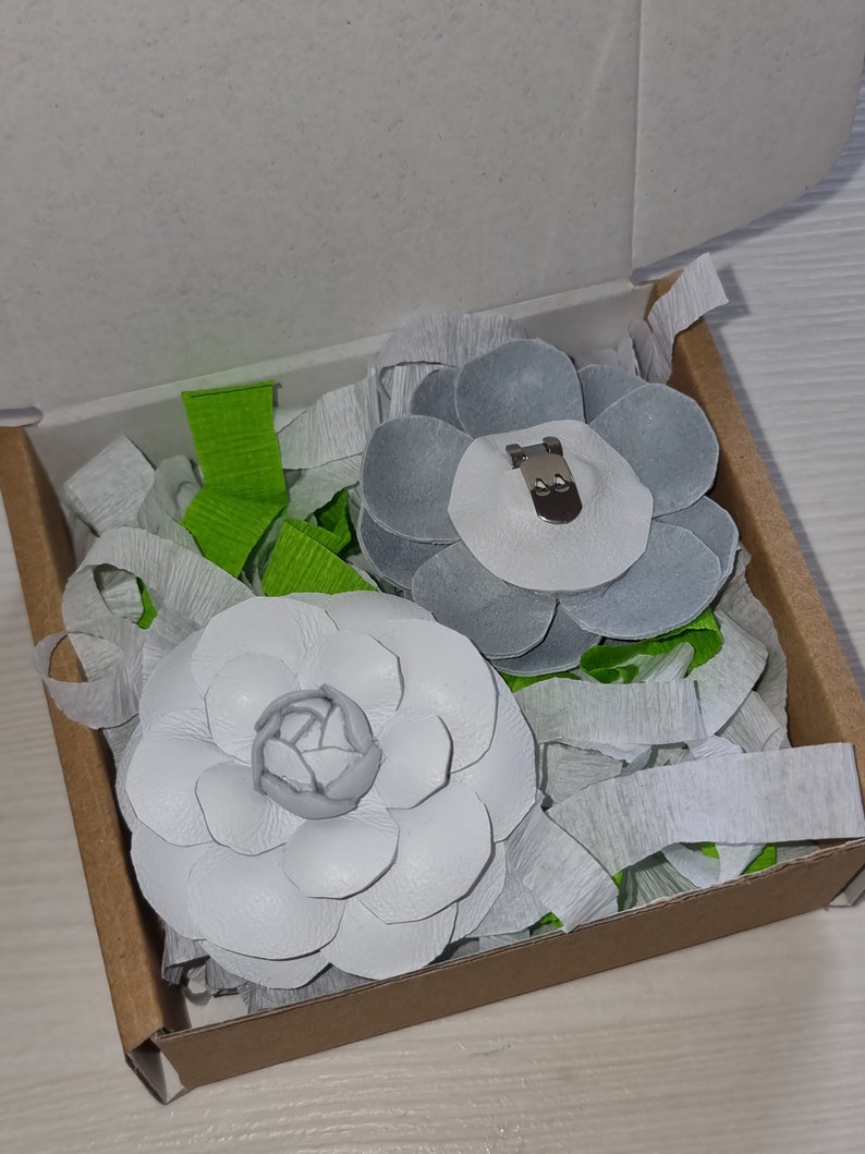 May include: Two white leather flower brooches in a cardboard box with green and white tissue paper. One brooch has a silver clip and the other has a white center.