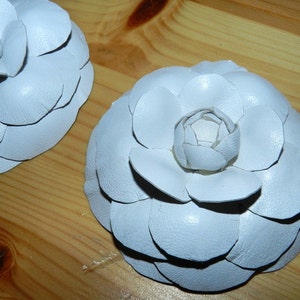 May include: Two white leather camellia flower brooches. The flowers are made of overlapping petals and have a slightly textured surface.