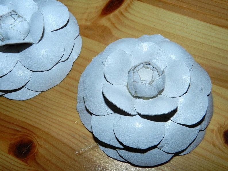 May include: Two white leather camellia flower brooches. The flowers are made of overlapping petals and have a slightly textured surface.