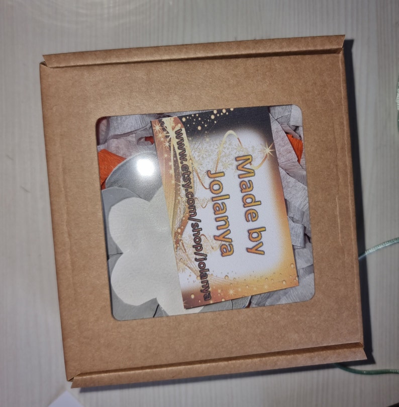 May include: A brown cardboard box with a clear window showing white flower-shaped decorations and a small card that says "Made by Jolanya" and "www.etsy.com/shop/jolanya".