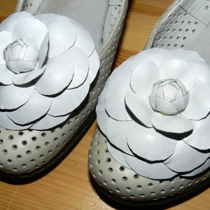 May include: A pair of white leather shoes with white flower embellishments. The shoes have a perforated design and are made of a soft, supple leather.