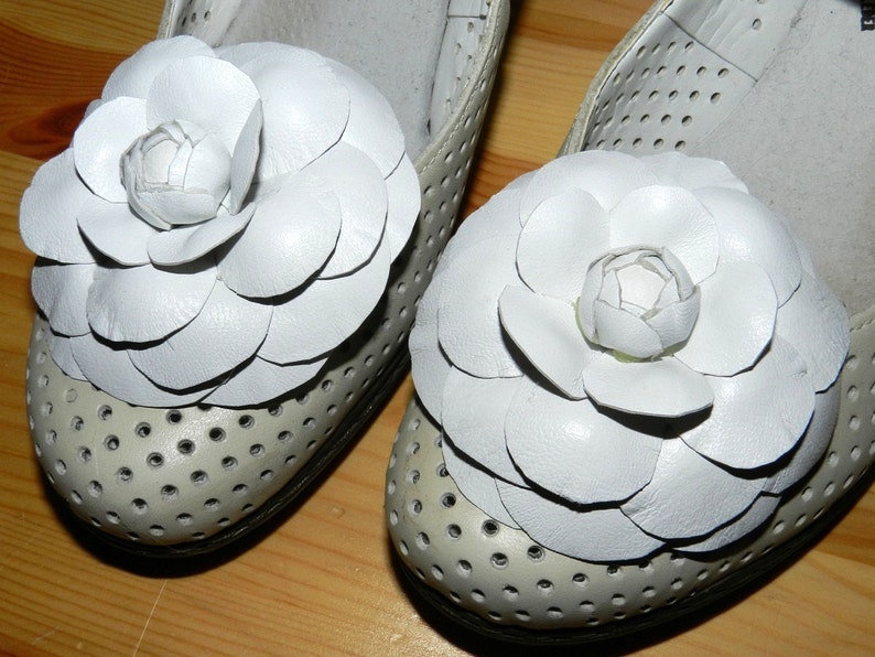 May include: A pair of white leather shoes with white flower embellishments. The shoes have a perforated design and are made of a soft, supple leather.