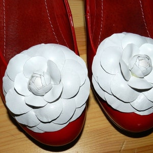 May include: A pair of red leather flats with white flower embellishments. The flowers are made of fabric and have a layered, petal-like design.
