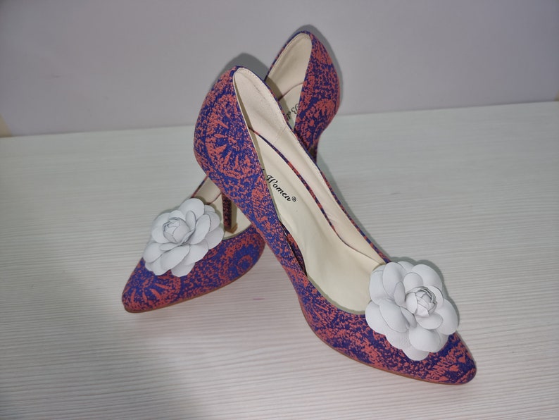 May include: A pair of high-heeled shoes with a floral pattern in shades of pink and blue. The shoes have a white flower embellishment on the toe.