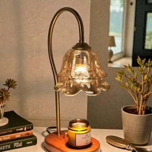 May include: A decorative table lamp with a flower-shaped glass shade in amber. The lamp has a curved brass stem and sits on a wooden base. A lit candle in a glass jar is on the base. A small potted plant and books are in the background.