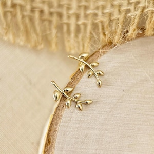 May include: A pair of gold-toned metal stud earrings, each shaped like a delicate branch with four small leaves. The earrings feature a polished, reflective surface and are designed to follow the curve of the earlobe when worn.