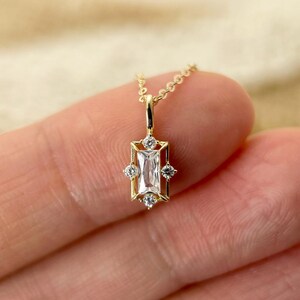 May include: A gold pendant necklace featuring a central rectangular baguette-cut clear stone framed by a geometric gold setting with four small round clear stones at the top, bottom, and sides, suspended from a delicate gold chain.