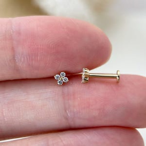 Op de afbeelding: Een gouden oorsteker met een platte achterkant en een bloemmotief met vier blaadjes, bestaande uit vier kleine, heldere, rondgeslepen steentjes. De steker heeft een dunne, rechte stift die aan de achterkant van de bloemvorm is bevestigd.