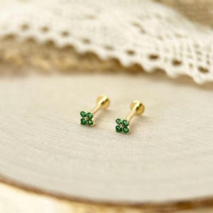 May include: A pair of gold stud earrings featuring a four-leaf clover design. Each earring is set with four small, round, vibrant green gemstones arranged in a cluster at the front, attached to a thin gold post with a flat circular backing.