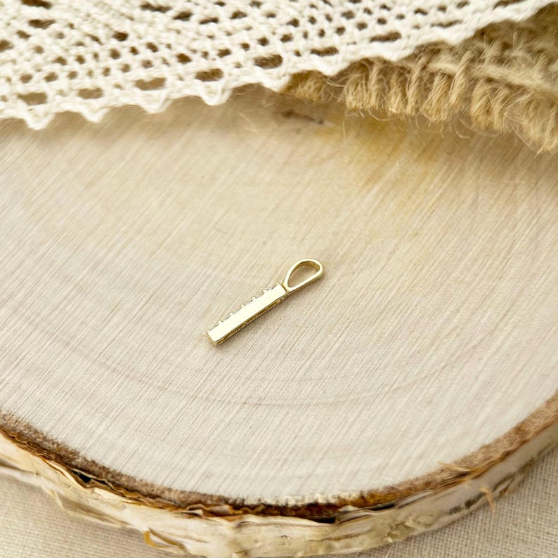 May include: A small, slender gold pendant featuring a straight, rectangular bar design with a series of tiny, clear inset stones along the front edge and a teardrop-shaped bail at the top for attachment to a chain or necklace.