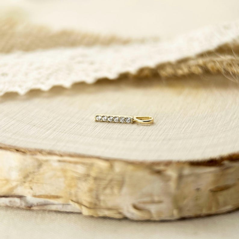 May include: A gold pendant featuring a thin, horizontal bar design set with five small, clear, round-cut stones. The pendant includes a small, oval-shaped bail at one end for attachment to a chain, showcasing a polished metallic finish throughout.