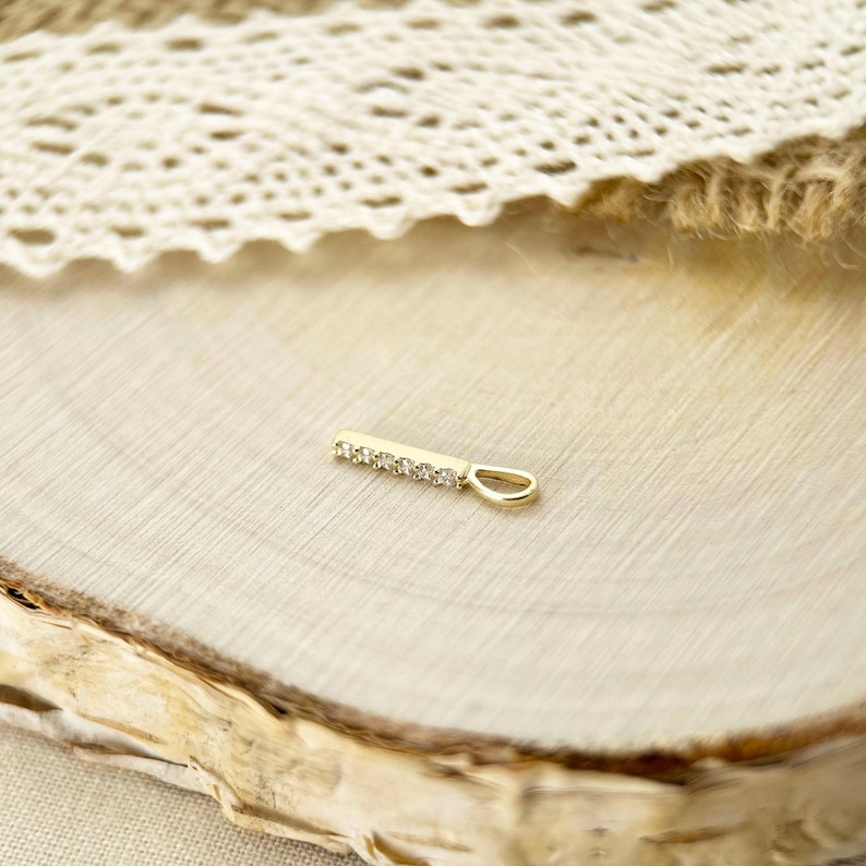 May include: A gold bar pendant featuring a row of five small, clear, round-cut stones set along the front surface. The pendant includes a small, oval-shaped bail at one end for attachment to a chain, showcasing a polished metallic finish.