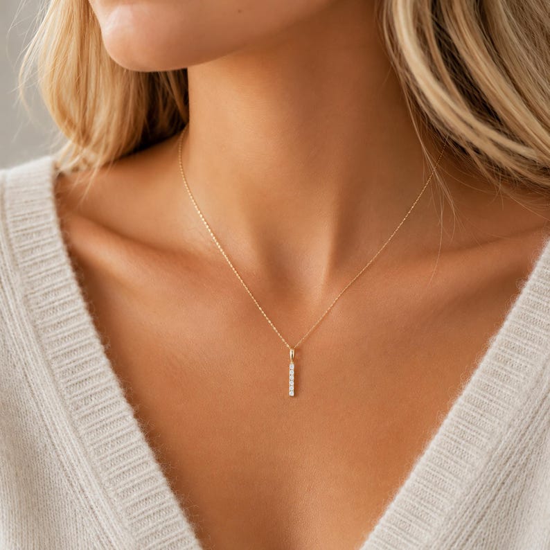 May include: A delicate gold chain necklace featuring a vertical bar pendant. The pendant is set with five small, clear, rectangular-cut stones arranged in a straight line, suspended from a thin, polished gold metal chain that rests on the collarbone.