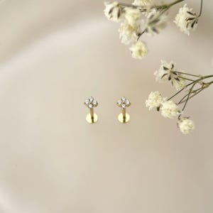 Puede incluir: Un par de pendientes de bot&oacute;n dorados, cada uno con un dise&ntilde;o de flor de cuatro p&eacute;talos engastado con peque&ntilde;as piedras redondas y transparentes. Los pendientes tienen un poste fino y recto y un cierre circular plano, junto a flores blancas.