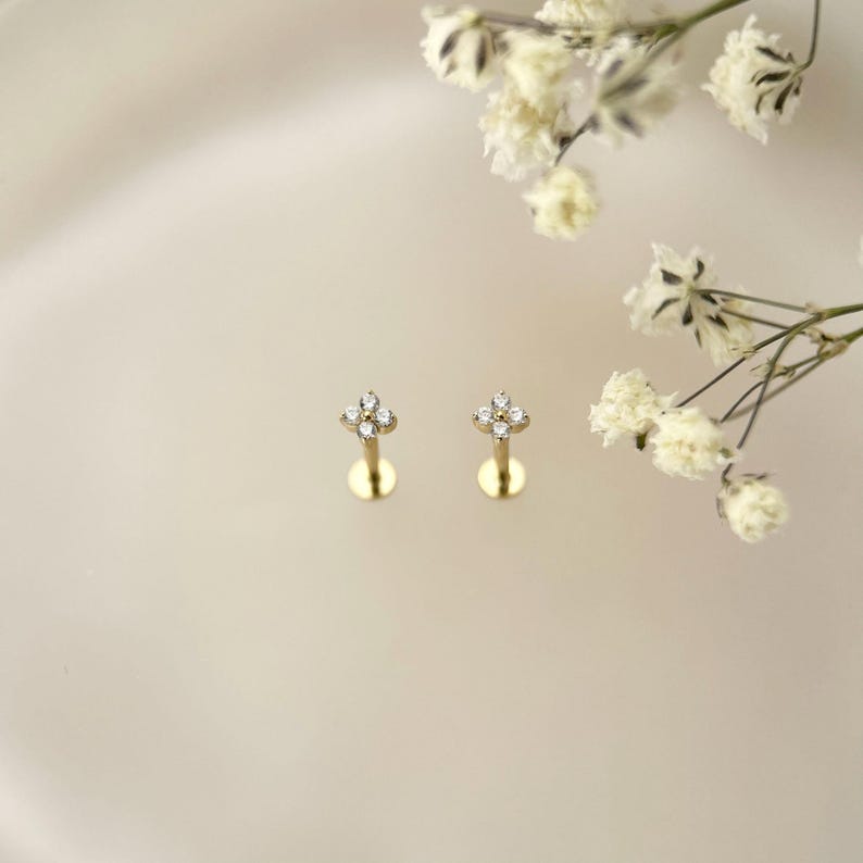 Puede incluir: Un par de pendientes de bot&oacute;n dorados, cada uno con un dise&ntilde;o de flor de cuatro p&eacute;talos engastado con peque&ntilde;as piedras redondas y transparentes. Los pendientes tienen un poste fino y recto y un cierre circular plano, junto a flores blancas.