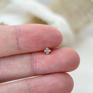 Puede incluir: Un peque&ntilde;o pendiente de bot&oacute;n dorado con un dise&ntilde;o de flor de cuatro p&eacute;talos, donde cada p&eacute;talo est&aacute; compuesto por una piedra preciosa transparente de corte redondo. El centro de la flor es una peque&ntilde;a cuenta dorada, creando una forma sim&eacute;trica.