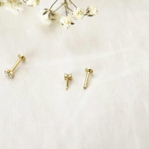 Puede incluir: Un pendiente tipo labret de color dorado con parte posterior plana y un peque&ntilde;o grupo de piedras transparentes en la parte superior. La pieza se muestra en tres partes: un pendiente ensamblado, un adorno con rosca y una base hueca plana.