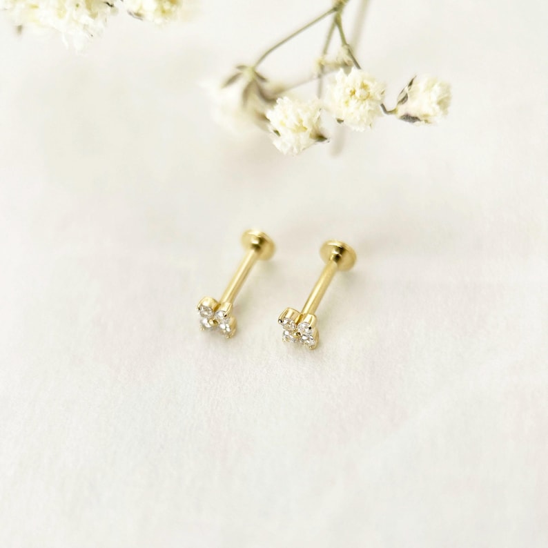 Puede incluir: Un par de pendientes de oro con engaste de cuatro garras, que presentan un grupo de cuatro peque&ntilde;as piedras transparentes dispuestas en forma cuadrada en la cabeza de cada poste, junto a peque&ntilde;as flores blancas.
