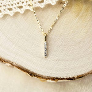 May include: A gold chain necklace featuring a vertical bar pendant. The slender bar is set with five small, clear, round-cut stones arranged in a straight line. The metal has a polished finish, and the chain links are delicate and uniform in size.