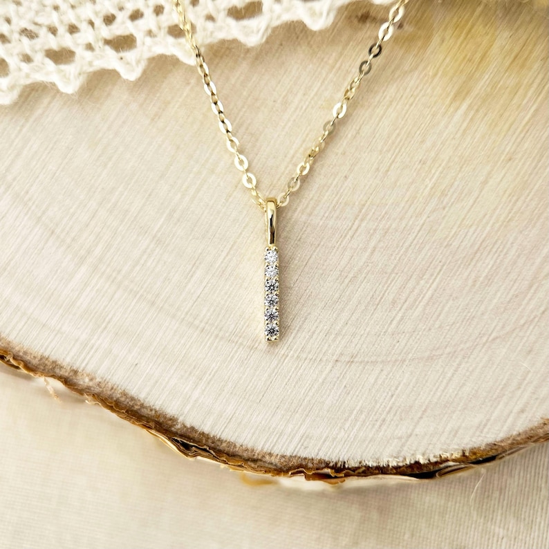 May include: A gold chain necklace featuring a vertical bar pendant. The slender bar is set with five small, clear, round-cut stones arranged in a straight line. The metal has a polished finish, and the chain links are delicate and uniform in size.