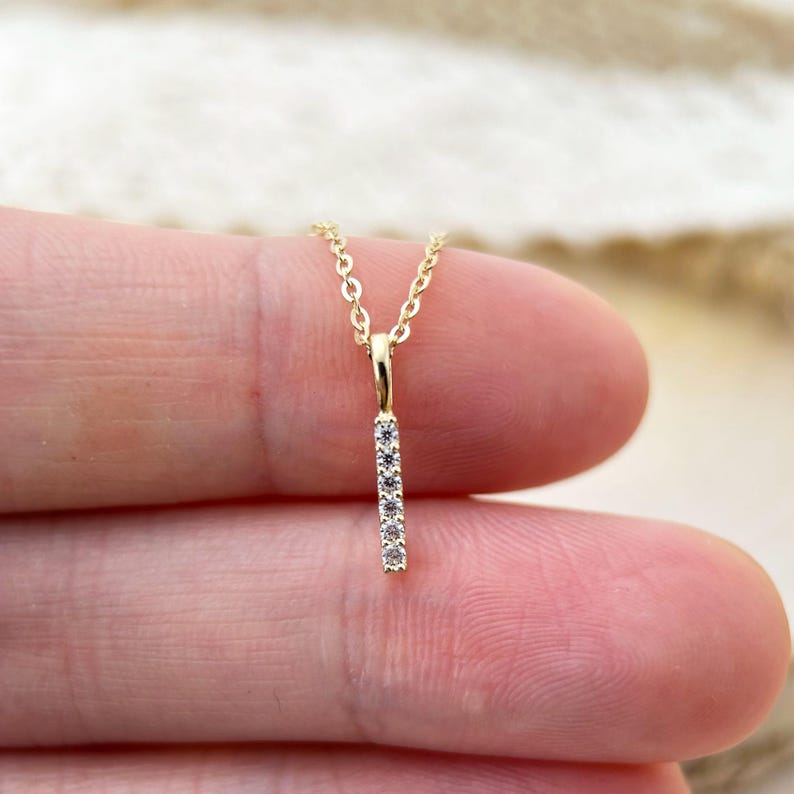 May include: A gold necklace featuring a vertical bar pendant set with five small, clear, round-cut stones. The pendant is attached to a delicate gold chain, creating a simple and refined piece of jewelry that hangs vertically from the bail.