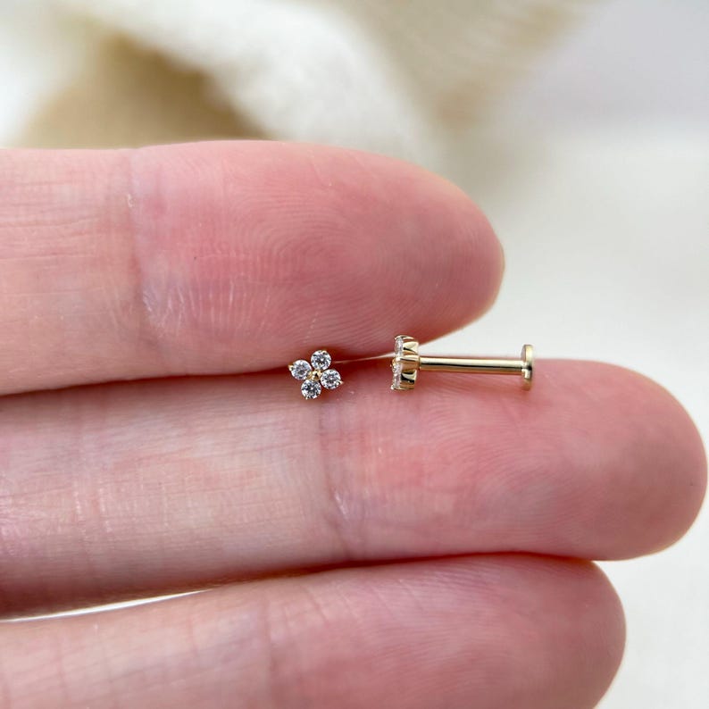 Puede incluir: Un pendiente de bot&oacute;n con parte posterior plana en tono dorado, con un dise&ntilde;o de flor de cuatro p&eacute;talos compuesto por cuatro peque&ntilde;as piedras transparentes de corte redondo. El pendiente tiene un poste recto y fino en la parte posterior.