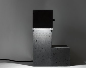 May include: A modern concrete table lamp with a black top and a light that shines through a clear panel. The lamp is on a white surface.