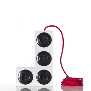 Op de afbeelding: Een witte L-vormige stekkerdoos met vier zwarte stopcontacten. Een rood elektriciteitskabel is op de vloer opgerold.
