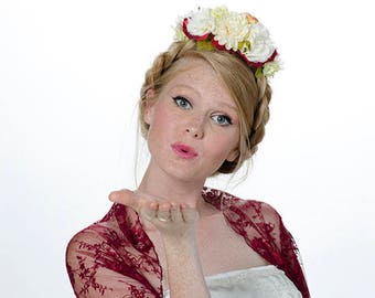 May include: A woman wearing a white dress with a burgundy lace shawl and a flower crown. She is blowing a kiss.