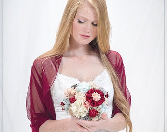 May include: A woman in a white dress with a burgundy shawl holds a bouquet of fabric flowers in shades of red, pink, cream, and blue.