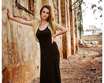 Può includere: Una donna con un vestito nero e un cardigan beige si trova di fronte a un muro di mattoni consumato dal tempo. Indossa una collana di perle e ha i capelli tirati indietro.