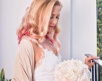 Può includere: Una sposa con i capelli biondi con riflessi rosa indossa un abito bianco di pizzo e uno scialle beige. Tiene un bouquet bianco di fiori di seta.