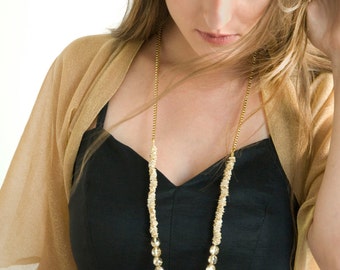Può includere: Una donna indossa un abito nero con una collana di perle dorate e uno scialle dorato trasparente.