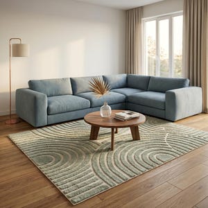 May include: A modern living room featuring a large, light blue sectional sofa, a round wooden coffee table, and a patterned rug in shades of green and beige. A floor lamp and window with curtains complete the scene.