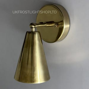 May include: A brass wall light with a conical shade and a circular backplate. The light is mounted on a white wall. The text "UKFROSTLIGHTSHOPLTD" is printed on the wall near the light.