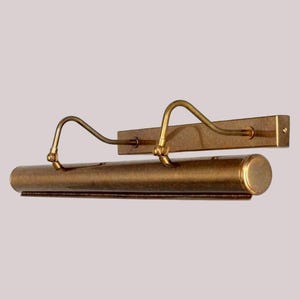 May include: A vintage-style picture light fixture in a warm brass finish. The fixture has a long, cylindrical light housing with two curved arms extending to the mounting plate. The design is classic and elegant.