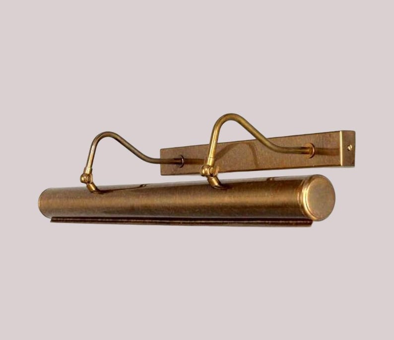 May include: A vintage-style picture light fixture in a warm brass finish. The fixture has a long, cylindrical light housing with two curved arms extending to the mounting plate. The design is classic and elegant.