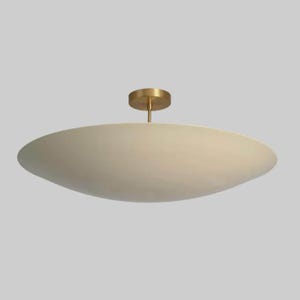 May include: A ceiling light fitting featuring a wide, shallow, matte off-white disc-shaped shade suspended from a central cylindrical brass-toned mount. The design is characterised by its clean, horizontal profile and smooth, uniform surface finish.