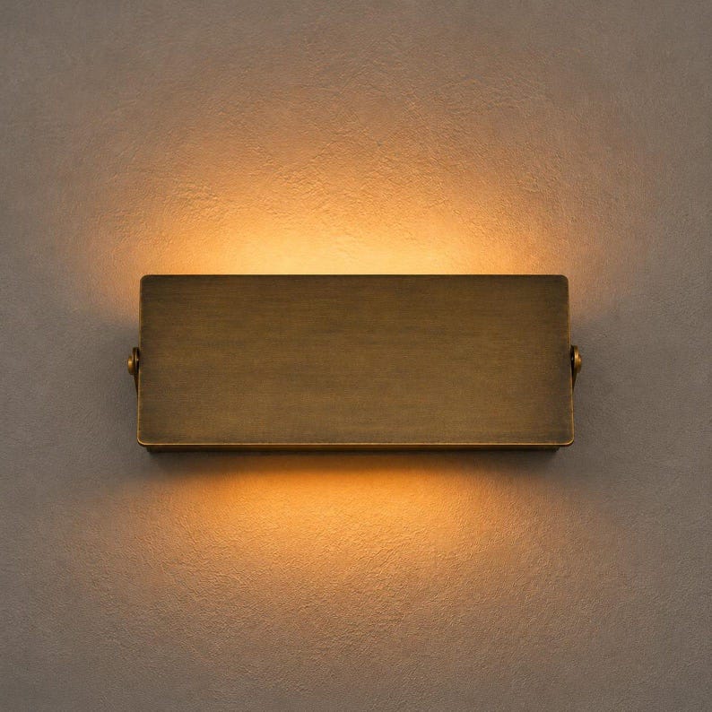 Op de afbeelding: Een rechthoekige wandlamp met een geborstelde antiek messing afwerking. Het armatuur heeft een massief metalen voorplaat met kleine ronde scharnieren aan de zijkanten. Het straalt warm licht uit aan zowel de boven- als onderkant.