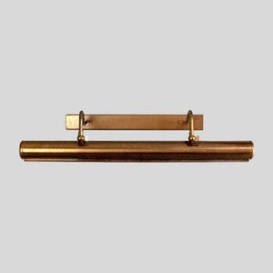 May include: A brass-toned picture light fixture. The fixture has a long, cylindrical light housing with a rectangular mounting plate. Two curved supports connect the light housing to the mounting plate. The light is designed to illuminate artwork.