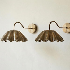 May include: Two brass wall sconces with a ruffled shade design. Each sconce features a curved arm and a circular wall mount. The shades have a scalloped edge, and the finish has a warm, antique appearance. Home decor.