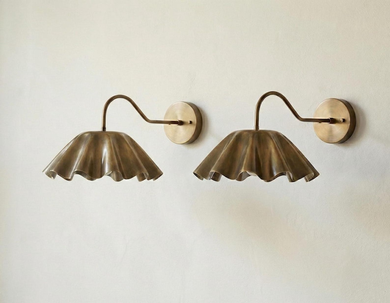 May include: Two brass wall sconces with a ruffled shade design. Each sconce features a curved arm and a circular wall mount. The shades have a scalloped edge, and the finish has a warm, antique appearance. Home decor.