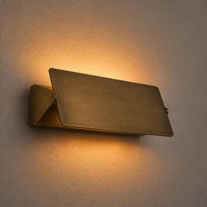 Op de afbeelding: Een rechthoekige wandlamp van geborsteld messing met een scharnierend voorpaneel. Het armatuur verspreidt een warme, zachte gloed van achter het paneel, waardoor het licht naar boven en beneden tegen de muur wordt geworpen voor diffuse verlichting.