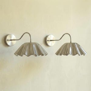 May include: Two brushed nickel wall sconces. Each sconce features a circular wall mount, a curved arm, and a ruffled, scalloped shade. The shades have a soft, textured appearance. These fixtures are designed for indoor lighting.