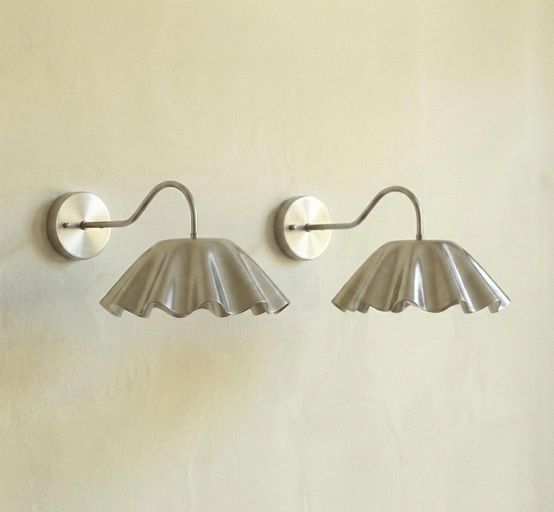 May include: Two brushed nickel wall sconces. Each sconce features a circular wall mount, a curved arm, and a ruffled, scalloped shade. The shades have a soft, textured appearance. These fixtures are designed for indoor lighting.