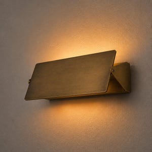 Op de afbeelding: Een rechthoekige wandlamp met een geborstelde messing afwerking, voorzien van een scharnierend voorpaneel dat naar buiten kantelt. Het armatuur verspreidt een warme, diffuse gloed vanaf de boven- en onderrand op de muur erachter.